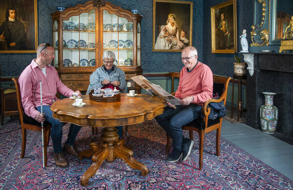 Drie bezoekers wagen zich aan een breiwerkje, lezen de krant en drinken een kopje thee in de Tuinkamer. Een van de bezoekers heeft een visuele beperking.